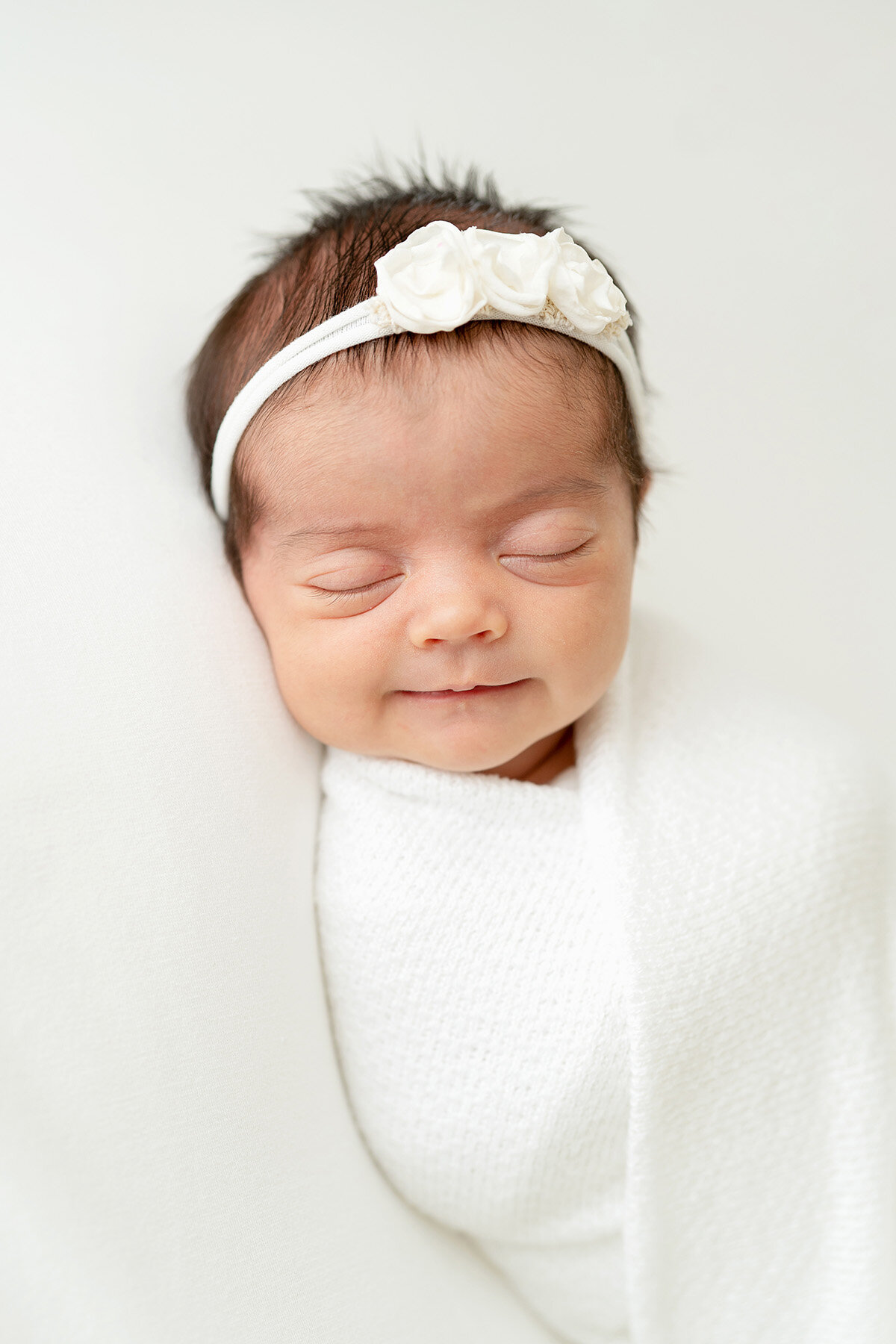smiling baby girl is swaddled in a white wrap and sleeping on a white blanket at Julie Brock Photography’s Louisville Ky studio