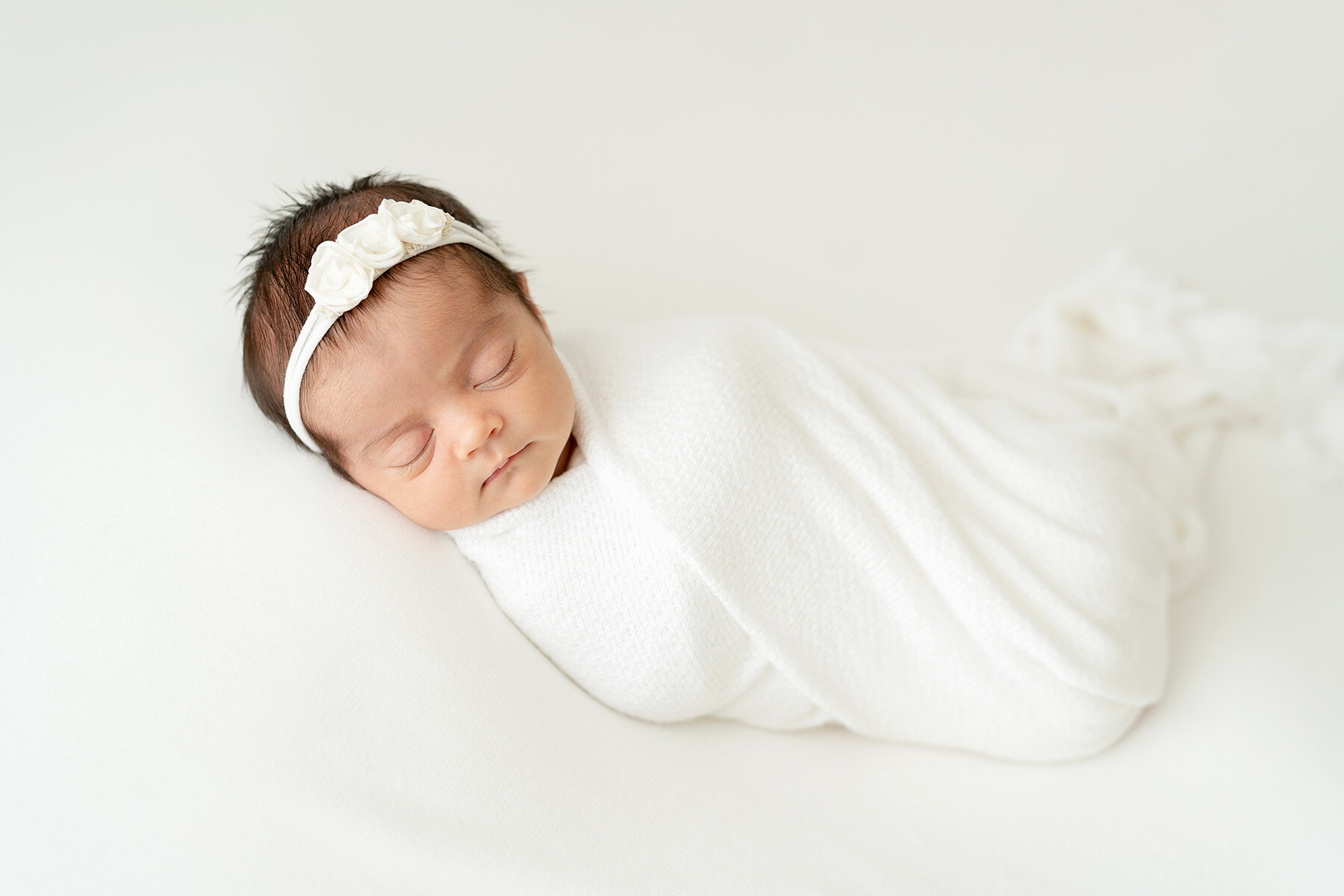 Simply styled white photography studio session. Sleeping baby is wrapped in white swaddle and sleeping on white blanket.