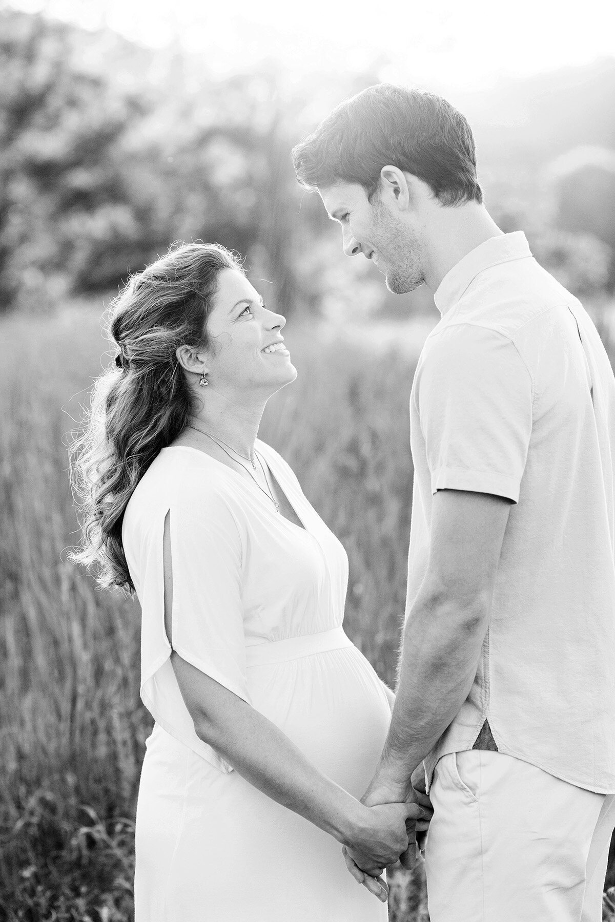 Maternity photo of a pregnant mother wearing  a white dress holding her husband’s hands in a Louisville KY field at sunset.