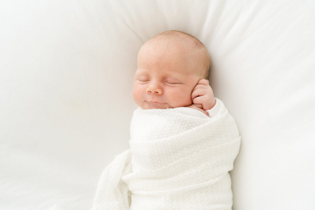 Newborn baby swaddled in a white blanket smiles during his photo shoot in Louisville Ky with Julie Brock, newborn photographer.
