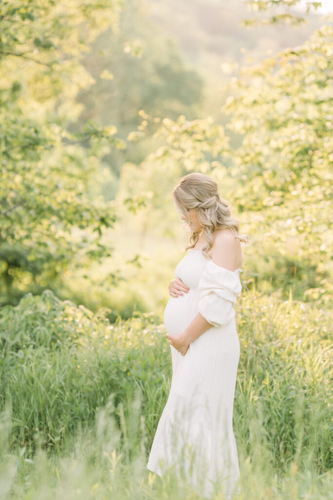 Louisville Ky mother wears off shoulder white dress and looks down at her baby bump during outdoor maternity photos with Julie Brock Photography