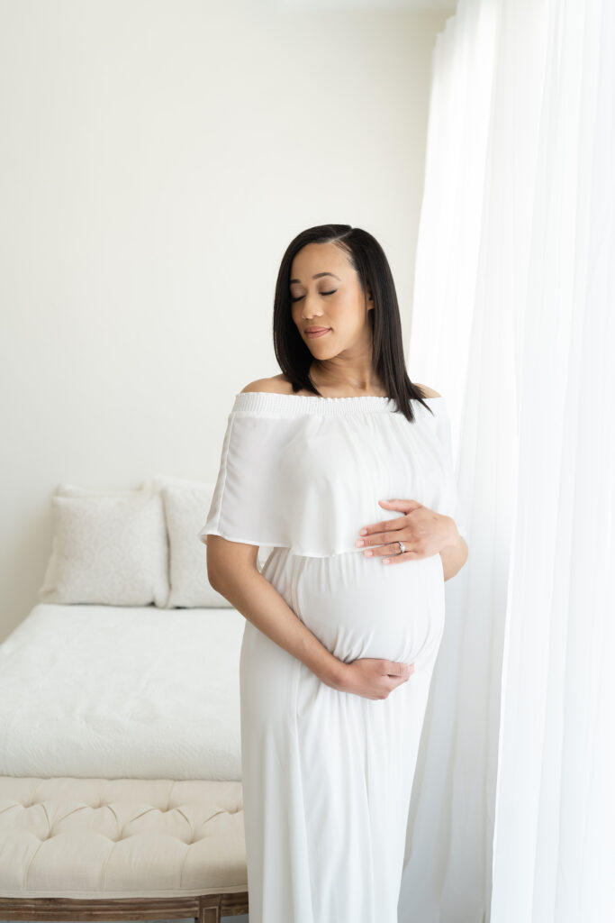 Pregnant mother wears off-shoulder-white dress and holds her belly at Julie Brock Photography in Louisville KY