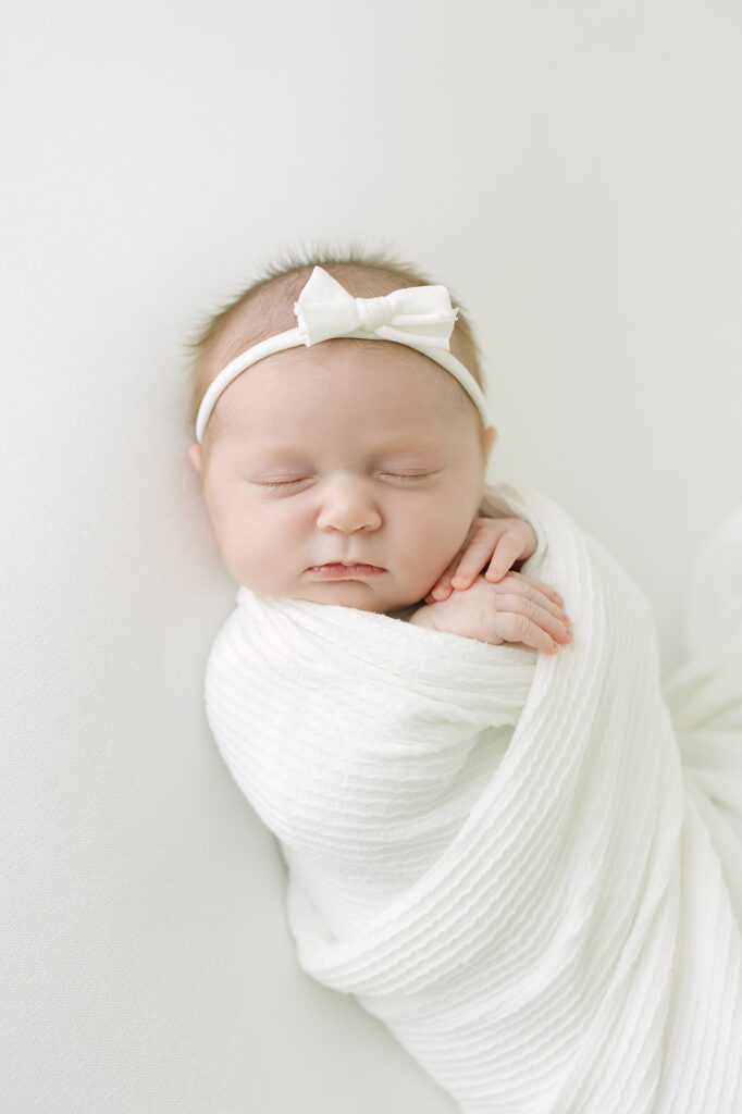 Louisville newborn photographer swaddles baby girl in white blanket and places white bow on her head