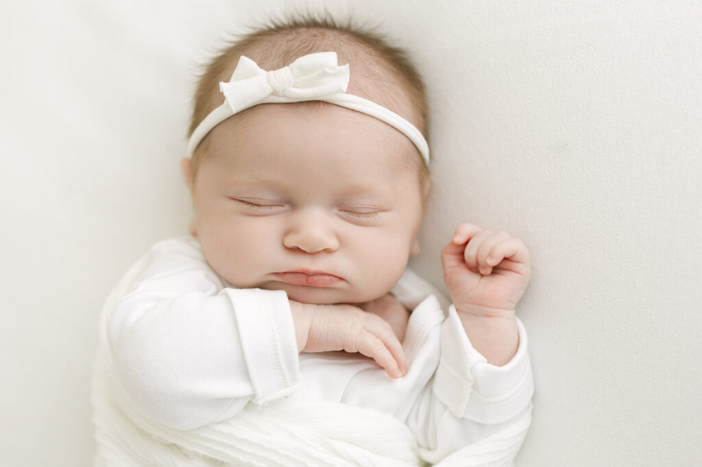 Sleeping baby girl wearing white onsie and bow at during Louisville newborn photography session.