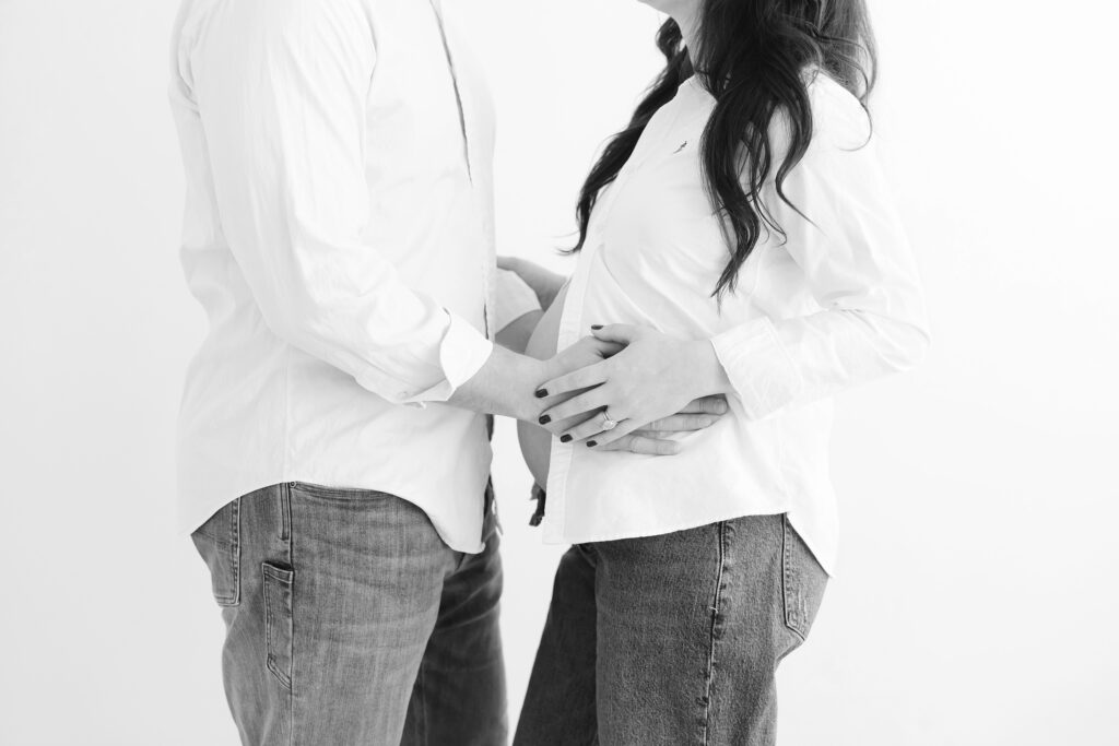 Up-close silhouette of hands placed on pregnant mother's growing baby bump. Parents are wearing white shirts and jeans at Julie Brock Photography in Louisville KY for their Maternity Session.  