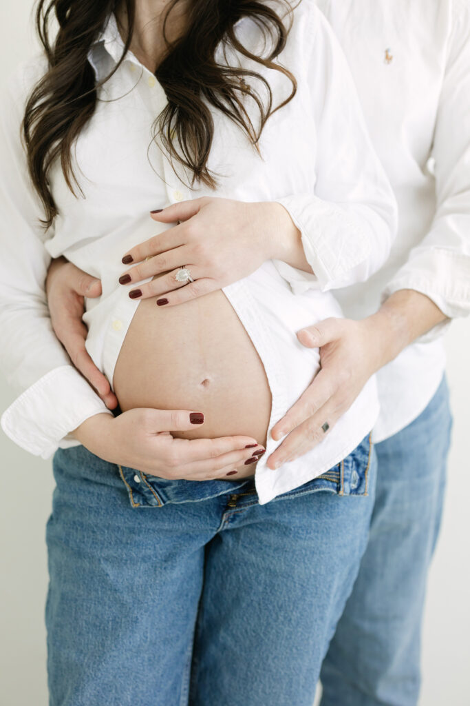 Expectant parents wearing white shirts and jeans wrap their arms around mom's pregnancy bump during their Louisville KY Maternity Session at Julie Brock Photography Studio