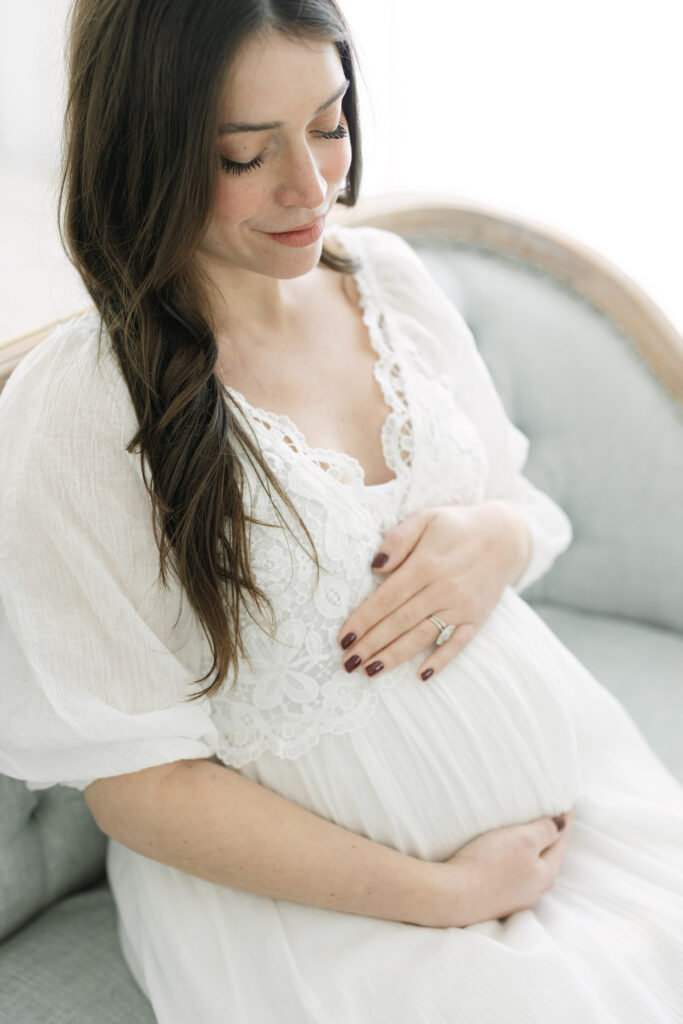 Pregnant mother wears delicate white dress while sitting on bench during a Maternity Session at Julie Brock Photography near Louisville KY