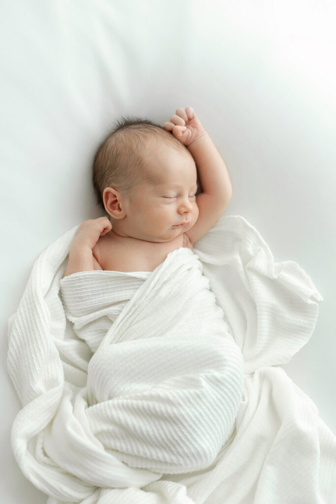 Newborn baby sleeps with arms stretched while wrapped in white knit blanket during a Newborn Session by Julie Brock Photography near New Albany Indiana and Louisville KY