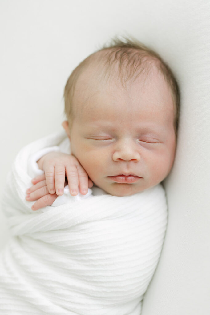 Sleeping baby boy is wrapped in soft white blanket with his hands tucked near his face during his Newborn Photo Shoot with Julie Brock Photography Studio near Louisville KY