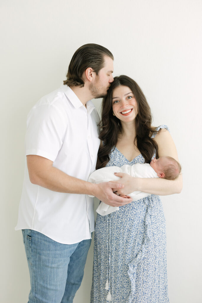 New parents hold their baby boy during their Newborn Photography Session near Louisville KY mom is wearing a blue summer dress dad is wearing a white shirt and jeans they're pose is relaxed and natural